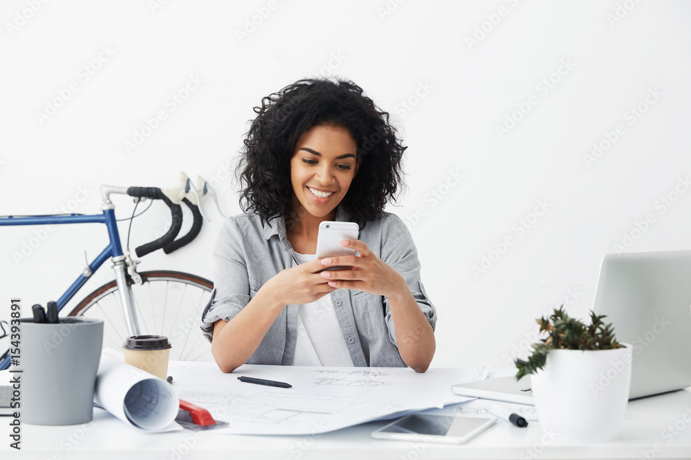 Wall mural Professional Afro American woman working as designer sitting in cosy office having happy expression while reading messages from her friends isolated over white background.People and technology concept