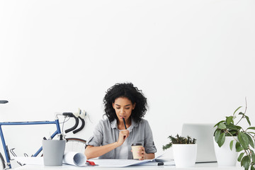 Fashionable female Afro American student doing her homework designing new buildings sitting in cosy home interior drinking coffee holding pen and having thoughtful expression. People and education