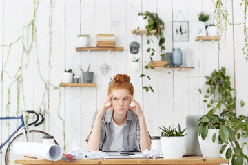 Portrait of Caucasian female architect sitting at her workplace over sketches, trying to focus on...