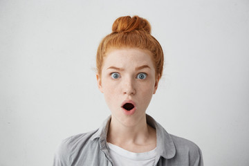 Close up shot of astonished freckled teenage girl wearing her ginger hair in bun raising eyebrows...
