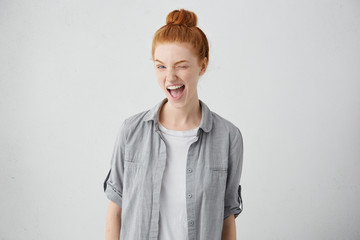 Childish and playful red haired teenage girl winking at camera, keeping mouth wide opened standing...