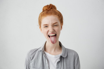 Naughty teenage girl with ginger hair and freckles misbehaving, sticking out her tongue at camera as a sign of disobedience, protest and disrespect. Human emotions, reactions, feelings and attitude © WHstudio Leushin N