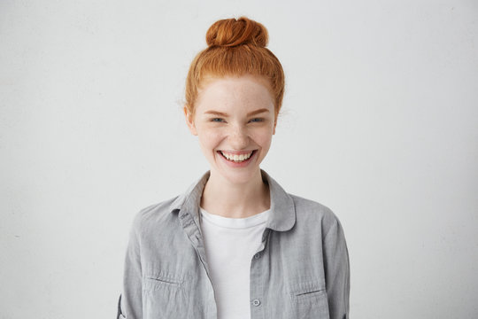 Beautiful ginger freckled girl with mysterious smile posing indoors at blank grey studio wall. Pretty woman with hair bun smiling broadly, showing white straight teeth, enjoying leisure time at home