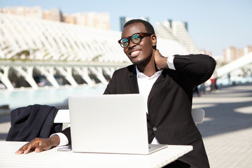 Young man with dark pure skin and white shining teeth realizing his future project while sitting outdoors over urban background using laptop pc touching his neck having some pain after hard work