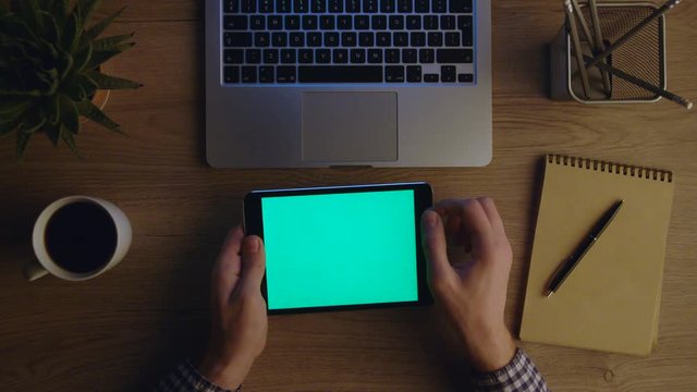 Man using tablet computer with green screen at office at night. Chroma key. Table top view with electronic devices