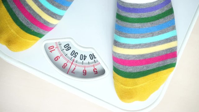 Female feet in colorful socks stepping on bathroom scales. Woman standing on weighing machine. Health care, weight loss concept. 4K ProRes HQ codec