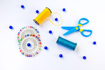 Fototapeta na wymiar Sewing spools of blue and yellow caprone threads, plastic scissors, glass beds and colorful pins on white background