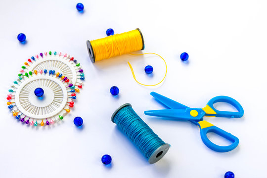 Sewing spools of blue and yellow caprone threads, plastic scissors, glass beds and colorful pins on white background