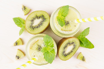 Fototapeta na wymiar Green kiwi fruit smoothie in glass jars with straw, mint leaf, cut ripe berry, close up, top view. White wooden board background.