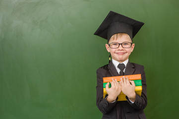 Boy in graduation cap with books standing near green chalkboard