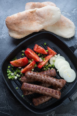 Barbecued balkan cevapcici served in a grill pan with onion, tomatoes and pita bread, above view