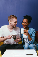 Happy and young interracial family at home on blue wall background, drinking morning coffee or tea. Togetherness, smile and happiness concept.