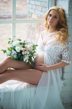 Beautiful pregnant girl smiling sitting on window with bouquet of flowers.