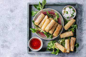 Fried spring rolls with red and white sauces, served in china plate on square wood tray with fresh green salad over gray blue texture background. Flat lay, space. Asian food - Powered by Adobe