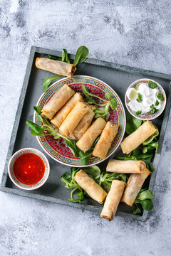 Fried spring rolls with red and white sauces, served in china plate on square wood tray with fresh green salad over gray blue texture background. Flat lay, space. Asian food