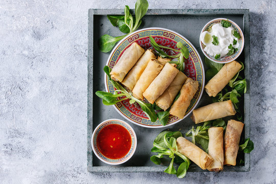 Fried spring rolls with red and white sauces, served in china plate on square wood tray with fresh green salad over gray blue texture background. Flat lay, space. Asian food