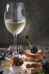 Grilled sandwich with melted goat cheese, blackberry, blueberry, rosemary and honey, served with glass of cold white wine over old metal texture background. Summer appetizer.