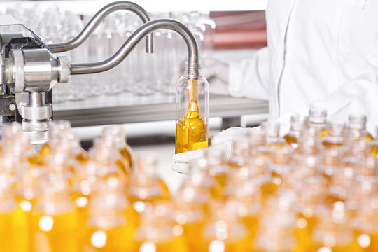 Professional scientist in white gown standing against factory background with many glass bottles filling them with thick yellow substance creating new lotion. Research, innovation, production concept