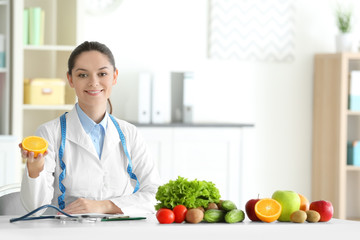 Young female nutritionist holding half of orange while sitting at table in her office