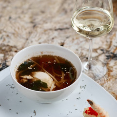 Japanese cuisine. Noodle soup udon in white ceramic bowl served with spoon, glass of white wine and king prawn on marble table