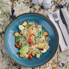 Cesar salad with baked chiken breast served in blue ceramic plate with glass of white wine, fork...
