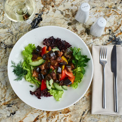 A warm salad of aubergines or eggplants and fresh vegetables served on white plate with glass of wine. Top view or flat lay.