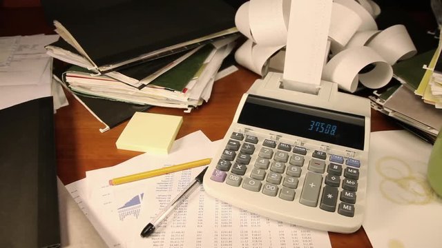 Camera dollies past a cluttered desk piled with paperwork.