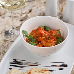Chicken fillet slices fried in spicy cream sauce with tortilla bread served in white bowl with glass of wine, knife and fork on napkin on marble table