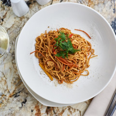 Flat egg noodles with vegetables served in big white ceramic round plate with glass of white wine on marble table