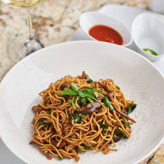 Flat egg noodles with duck meat, chicken, shiitake mushrooms served on white plate with white garlic and red spicy sauce