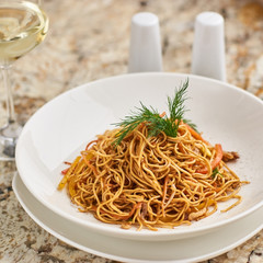 Thin egg noodles with vegetables and soy sauce served on white round plate with glass of white wine and chopsticks