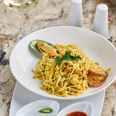 Flat egg noodles with seafood served in big white ceramic round plate with glass of white wine on marble table