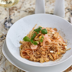Rice noodles with peanuts, chicken and shrimps served on white round plate with glass of white white on marble table