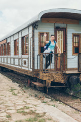 Obraz na płótnie Canvas Young woman waving on railway station. Beautiful girl in blue t-shirt with backpack standing in old train wagon. DaLat Railway Station, may 2017