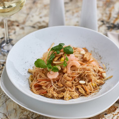 Rice noodles with peanuts, chicken and shrimps served on white round plate with glass of white white on marble table