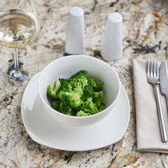 Steamed broccoli served in white bowl with glass of white wine on marble background