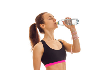 young fitness girl stands sideways in the sports top and drinking water from a bottle