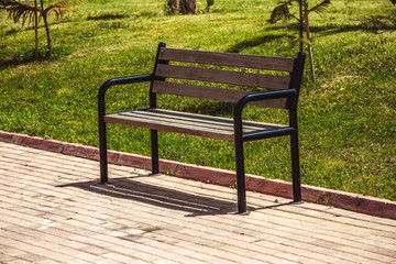 An empty wooden bench stands in the park on the sidewalk. On the background there is a green grass and lawn. No one. A clear summer day in nature.