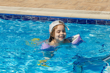 A charming child of European appearance, a girl 4-5 years old, swimming in the pool. Summer, a clear and sunny day. A girl with her family on vacation at sea. The girl has inflatable arm ruffles.