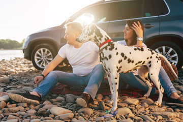 Portrait of happy young adult couple with dog on roadtrip. Man sitting on plaid with woman. Outdoor...