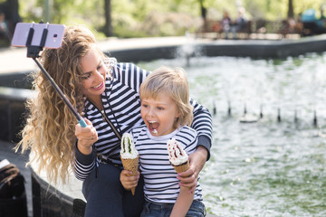 Fototapeta na wymiar Cute little boy and his mother staining each other with ice cream near the fountain outdoors. Young pretty mom and her son having fun together and making selfie