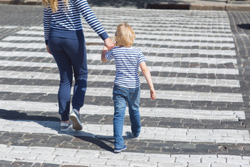 Young mother and her little child crossing the street holding by the hand. Boy and adult woman walking on the crosswalk on the road with hard traffic.