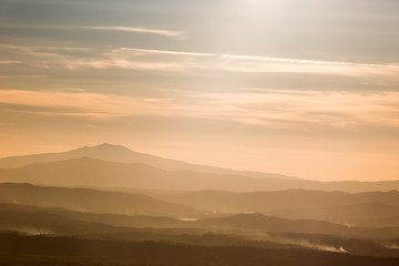 Various layers of mountains and hills in the middle of a golden, orange light during sunset, with clouds also reflecting light, and smoke on the right bottom corner