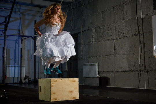 Beautiful and muscular blonde bride in a wedding dress jumping into wooden boxes in a training. Crossfit
