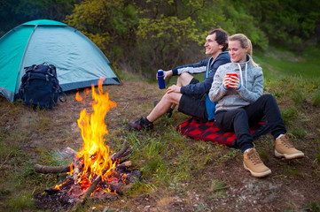 Romantic couple resting near bonfire with cup of tea, autumn forest background. Young blonde woman and handsome man. Camping, travel, tourism, hike and people concept.