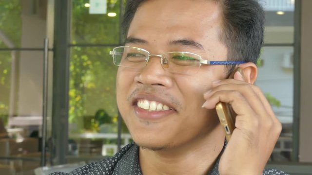 Close up of a young Asian male professional chatting with a colleague on his smartphone. Shot then pulls out and reframes to a medium close up