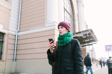 The girl on the street during a winter time is very puzzled with something. On her a lilac cap, a green scarf and a blue jacket. The girl holds phone in hand.