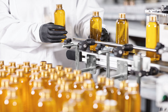 Research and development concept. Hands of professional worker of factory putting glass bottles with yellow liquid on conveyor line. Automated process of producing cosmetic products on factory