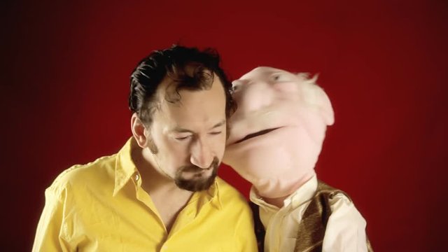 The puppet of an old man telling secrets to a funny ugly guy.
