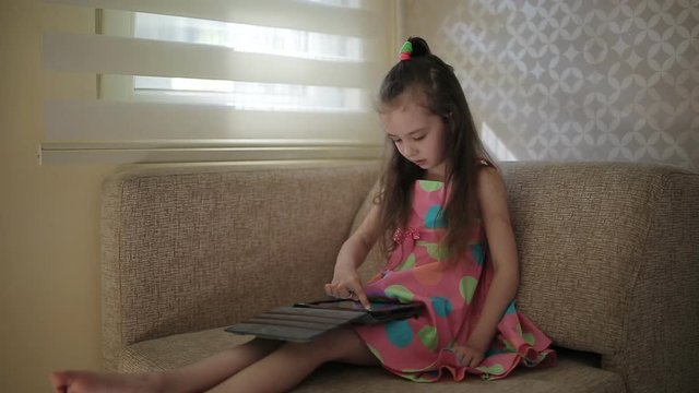 Little cute girl sitting on sofa and playing with tablet. Little girl in a beautiful colored dress sits on a chair by the window and uses a tablet